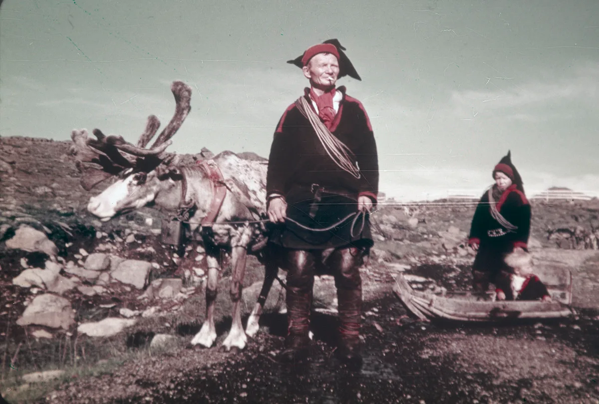 Sámi man in traditional clothing stands with reindeer in Arctic landscape, circa mid-20th century.
