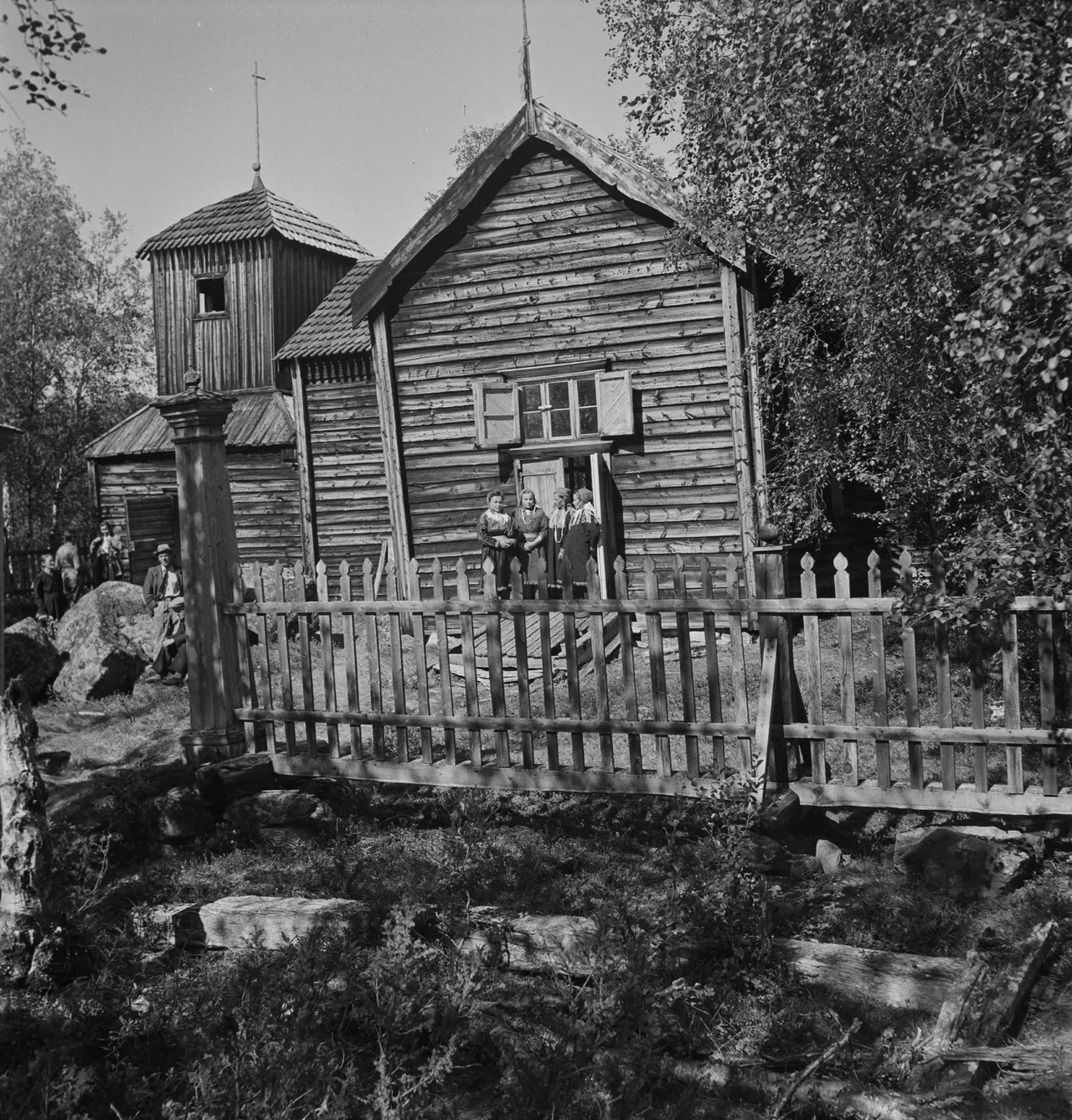 Traditional wooden Sámi chapel with bell tower surrounded by birch trees and visitors, Northern Europe.